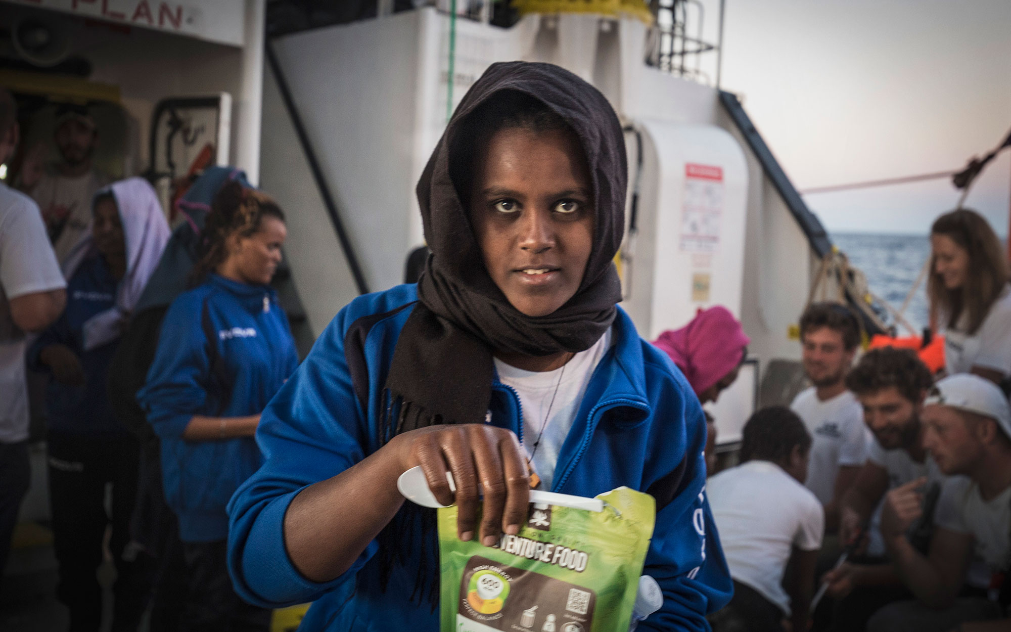 A woman holds a ready-to-eat meal packet as members of the SOS MEDITERRANEE crew chat with other rescued persons on board the Aquarius. October 2017.